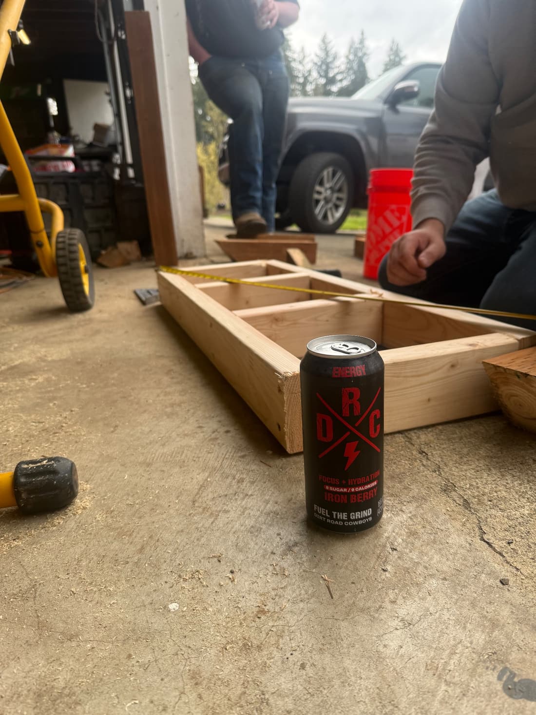 Iron Berry can on a concrete floor next to a wood frame and tape measure, with people working behind it