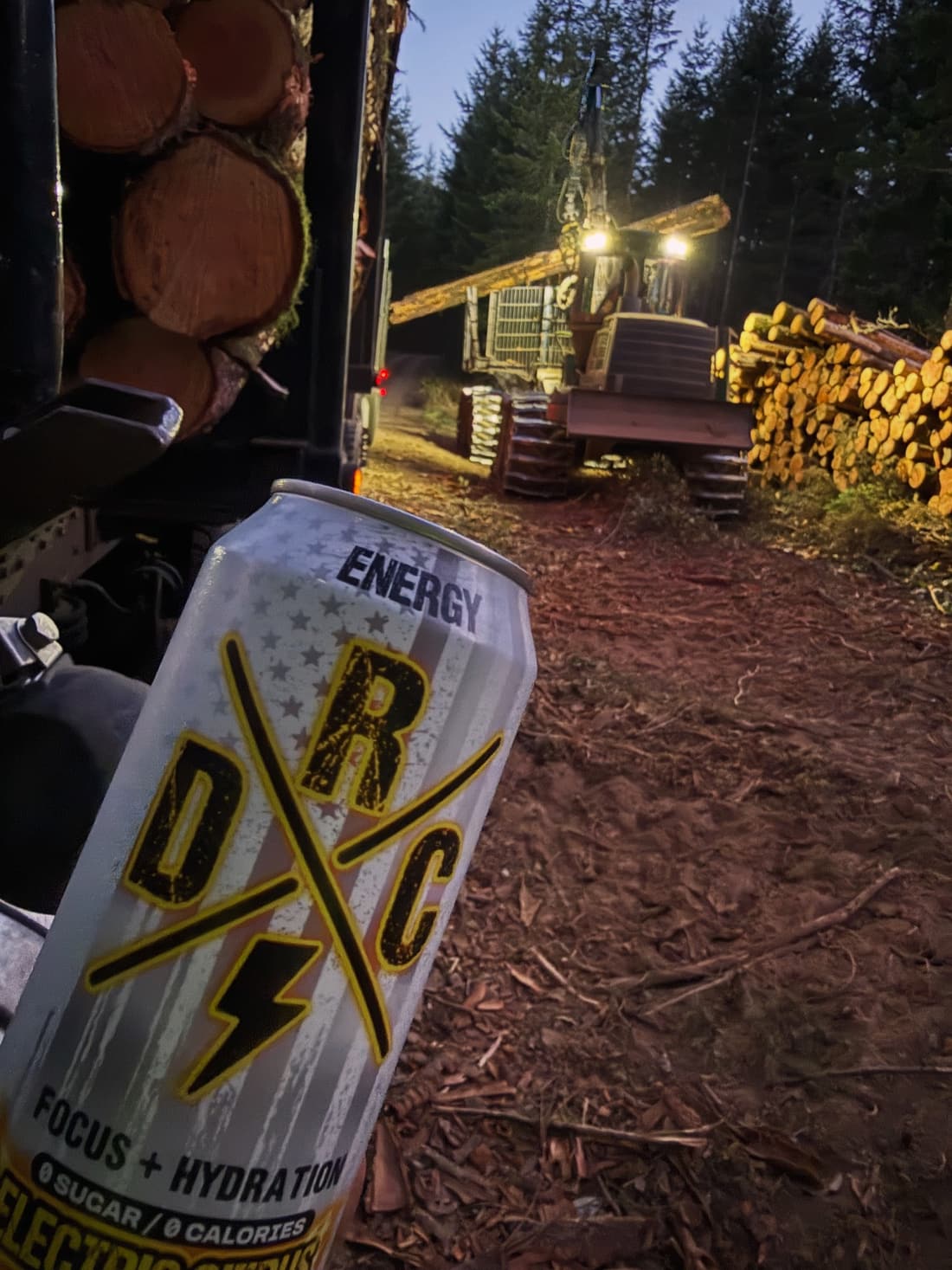 Electric Citrus can in the foreground with a logging truck load, a logging machine with its lights on and a log pile at dusk
